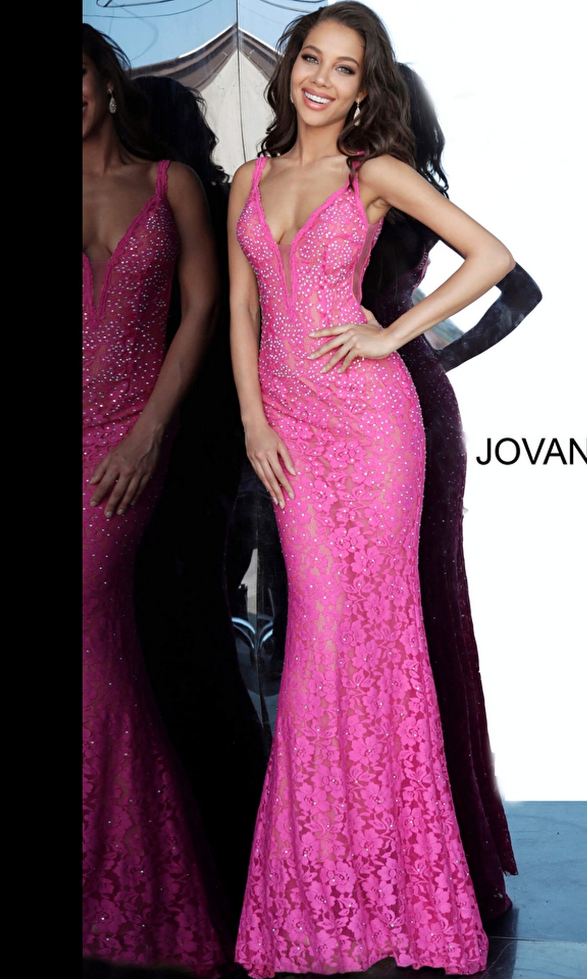 A woman in the Jovani 48994 Illusion-Sides Long Lace Prom Dress stands smiling in front of a reflective glass wall, showcasing the fitted, floor-length bright pink gown with rhinestone details.